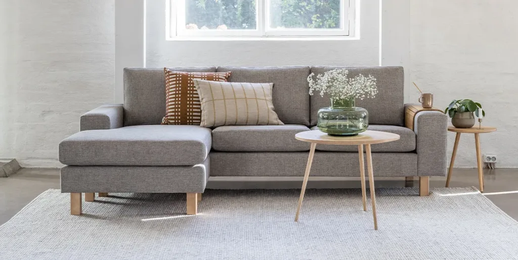 Beige Studio divaanisohva, jonka edessä sohvapöytä ja lasimaljakko jossa kukkia. Sohvan divaani on vasemmalla puolella edestä päin katsottuna.