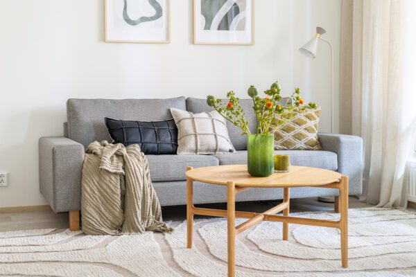 Kolmen istuttava Studio-sohva verhoiltuna vaaleanharmaalla kankaalla. Sohvalla tyynyjä ja viltti. Sohvan edessä puinen sohvapöytä, jonka päällä maljakko, jossa kukkia.