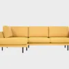 Kanerva-avokulmasohva verhoiltuna keltaisensävyisellä kankaalla ja jalkoina mustat pyöreät metallijalat, tuotekuva edestäpäin kuvattuna, jossa divaani vasemmalla.
