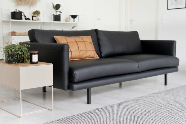Kolmen istuttava sohva verhoiltuna mustalla nahalla ja somisteena ruskea sisustustyyny, sohvassa mustat metallijalat, sisustuskuva.