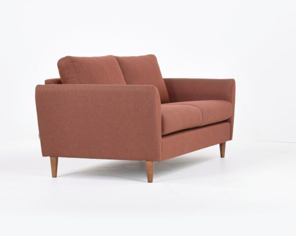 Kuura-2,5 istuttava sohva punertavalla kankaalla verhoiltuna ja jalkana pähkinä puujalka, tuotekuva viistosti kuvattuna.