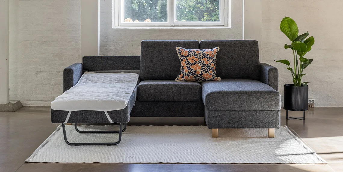 Tummanharmaa Aina divaanivuodesohva, jossa yksi vuode avattuna ja pedattuna kevyellä petauspatjalla. Divaani oikealla puolella edestä päin katsottuna.