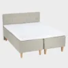 Vaalean beige 160 cm leveä ja 200 cm pitkä runkopatja napillisella sängynpäädyllä ja tammen värisillä kantikkailla puujaloilla.