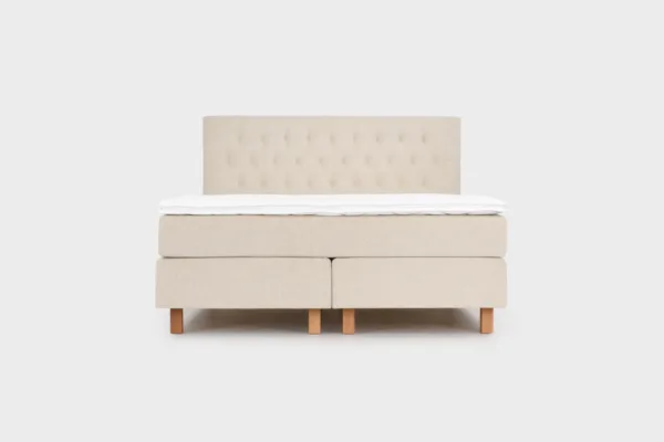 Vaalean beige jenkkisänky 180x210 cm tammen värisillä jaloilla ja vaalean beigellä sängynpäädyllä. Kuvattuna suoraan edestä päin vaaleanharmaata taustaa vasten.