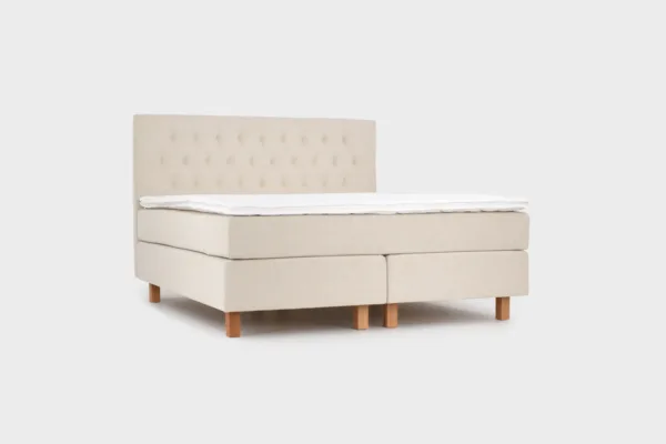 Vaalean beige jenkkisänky 180x210 cm tammen värisillä jaloilla ja vaalean beigellä sängynpäädyllä. Kuvattuna viistosti edestä päin vaaleanharmaata taustaa vasten.