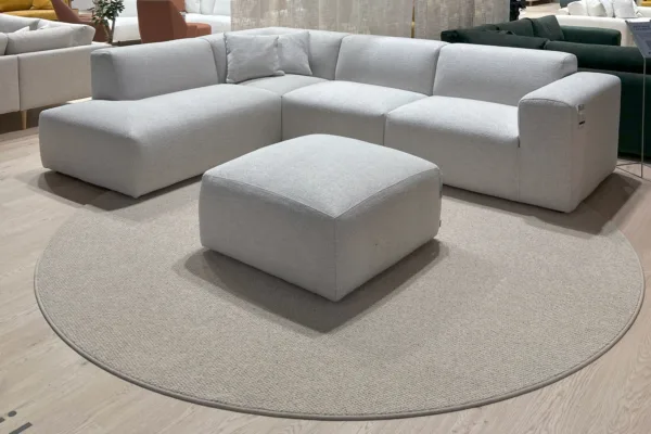 VM Carpetin matto Hehku greigen sävyssä laminaattilattian päällä ja avokulmasohvan sekä rahin kanssa kuvattuna messuympäristössä.