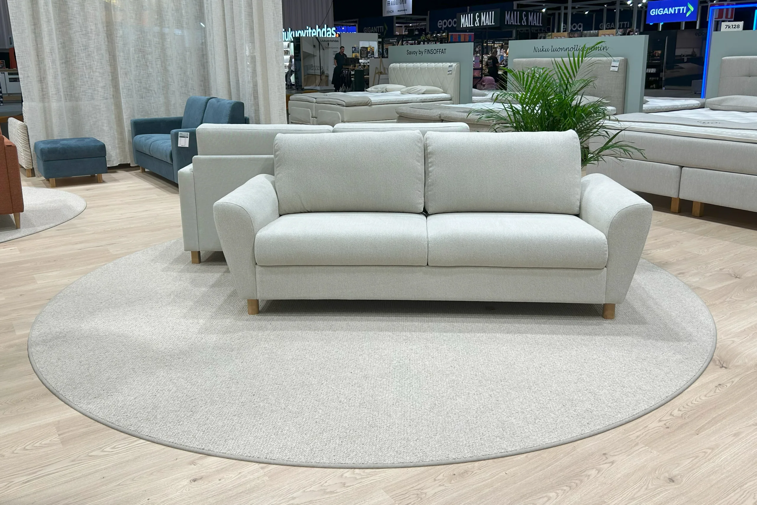 VM Carpetin matto Hehku greigen sävyssä laminaattilattian päällä valkoisen sohvan kanssa kuvattuna messuympäristössä.