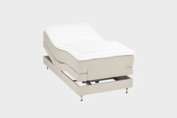 Vaalean beige moottorisänky 100x200 cm alumiinin värisillä jaloilla. Yhden hengen säätösänky on kuvattuna viistosti päädyt aavistuksen koholla vaaleaa taustaa vasten.