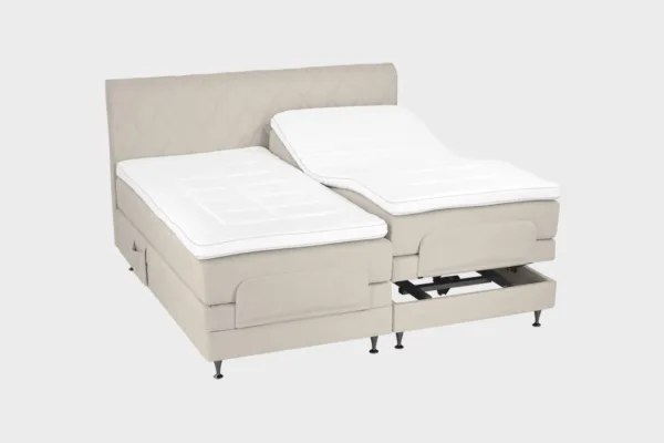 Vaalean beige moottorisänky koossa 180x200 cm mustilla jaloilla sekä samanvärisellä sängynpäädyllä. Parisänky on kuvattuna viistosti toisen sänkypuoliskon päädyt aavistuksen koholla vaaleaa taustaa vasten.